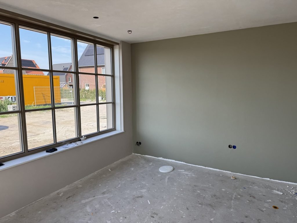 Nieuwbouwkamer met grote roedenramen en een groengrijs geverfde wand, buiten nieuwe rijtjeshuizen