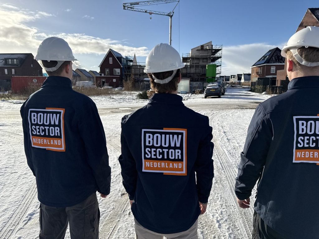 Drie vakmensen met witte helm en bedrijfsjas lopen door een besneeuwde nieuwbouwstraat met een bouwkraan